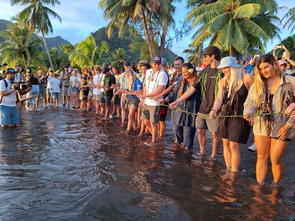 Surfeurs et officiels réunis sur la plage lors de la cérémonie de l’eau, une longue tresse de ‘autῙ dans les mains.